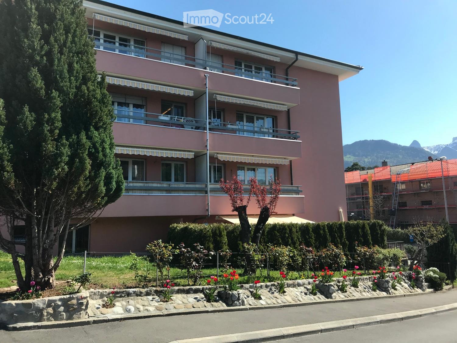 Two-story pink building with balconies, large windows, and striped awnings. Surrounded by a well-maintained garden with flowers and shrubs.