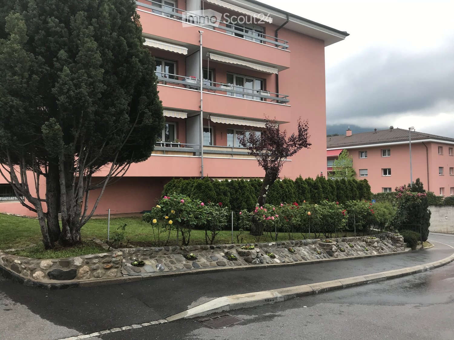 Pink multi-story apartment building with balconies, surrounded by greenery and flowers.