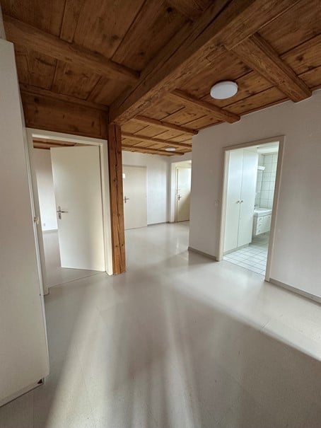 Empty room with wooden ceiling beams, white walls, and a smooth concrete floor. The space is completely unfurnished, with visible doorways leading to other rooms.