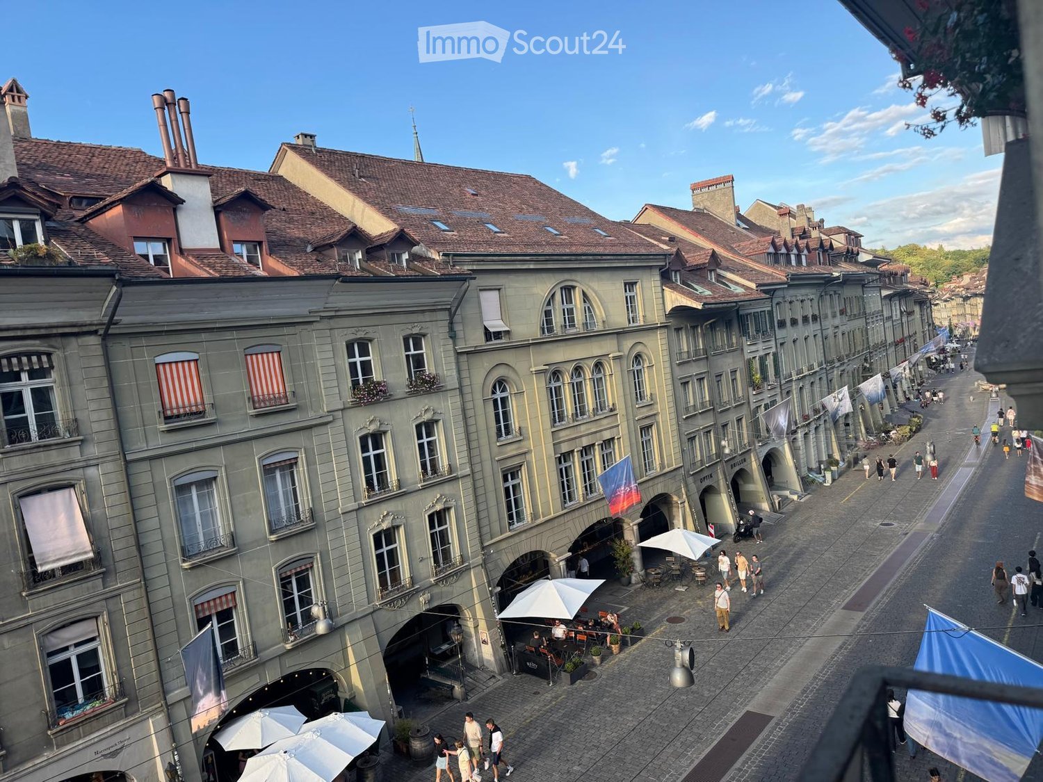 A historic European street lined with multi-story buildings featuring classic architecture, large windows, and ornate facades. The street is paved with cobblestones and has outdoor seating areas with umbrellas, people walking, and flags hanging from the b