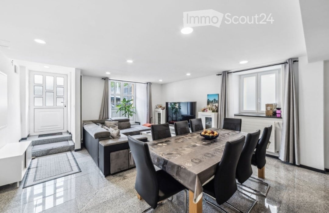 Open-plan living and dining area with a grey sectional sofa, coffee table, dining table with chairs, television, fireplace, and decorative plant.