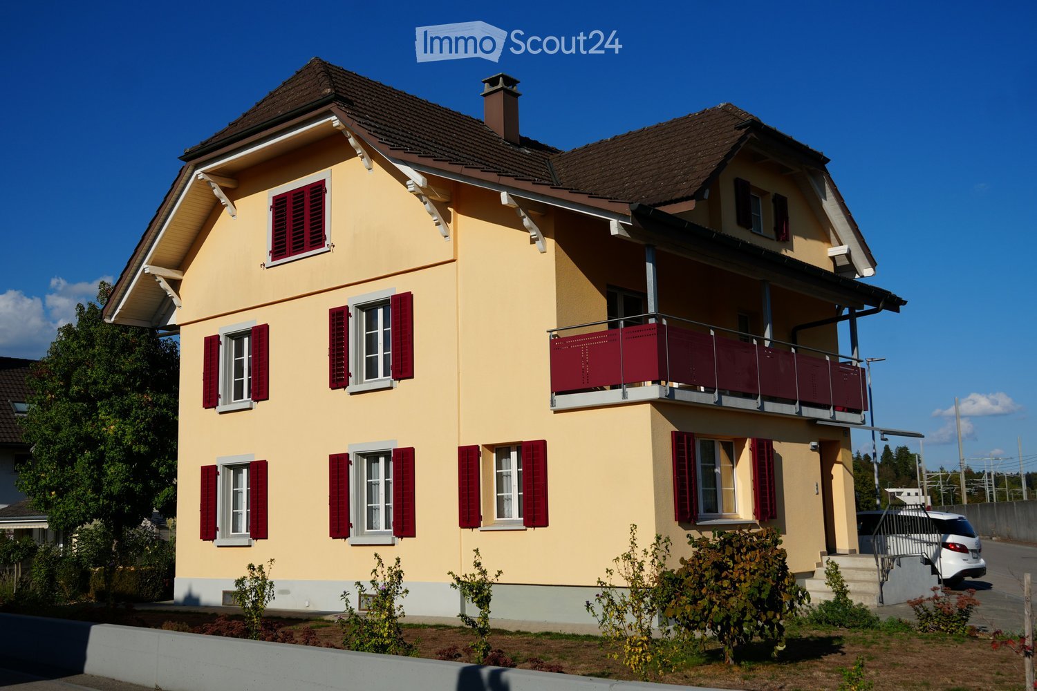 Two-story house with yellow exterior, red shutters, and a balcony on the upper floor. The house has a brown tiled roof and is surrounded by small shrubs and a driveway with a parked car.