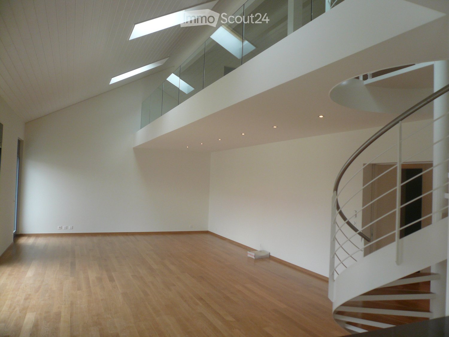 Empty room with wooden flooring, white walls, and a modern spiral staircase with a glass railing. The room features recessed lighting and a large skylight above.