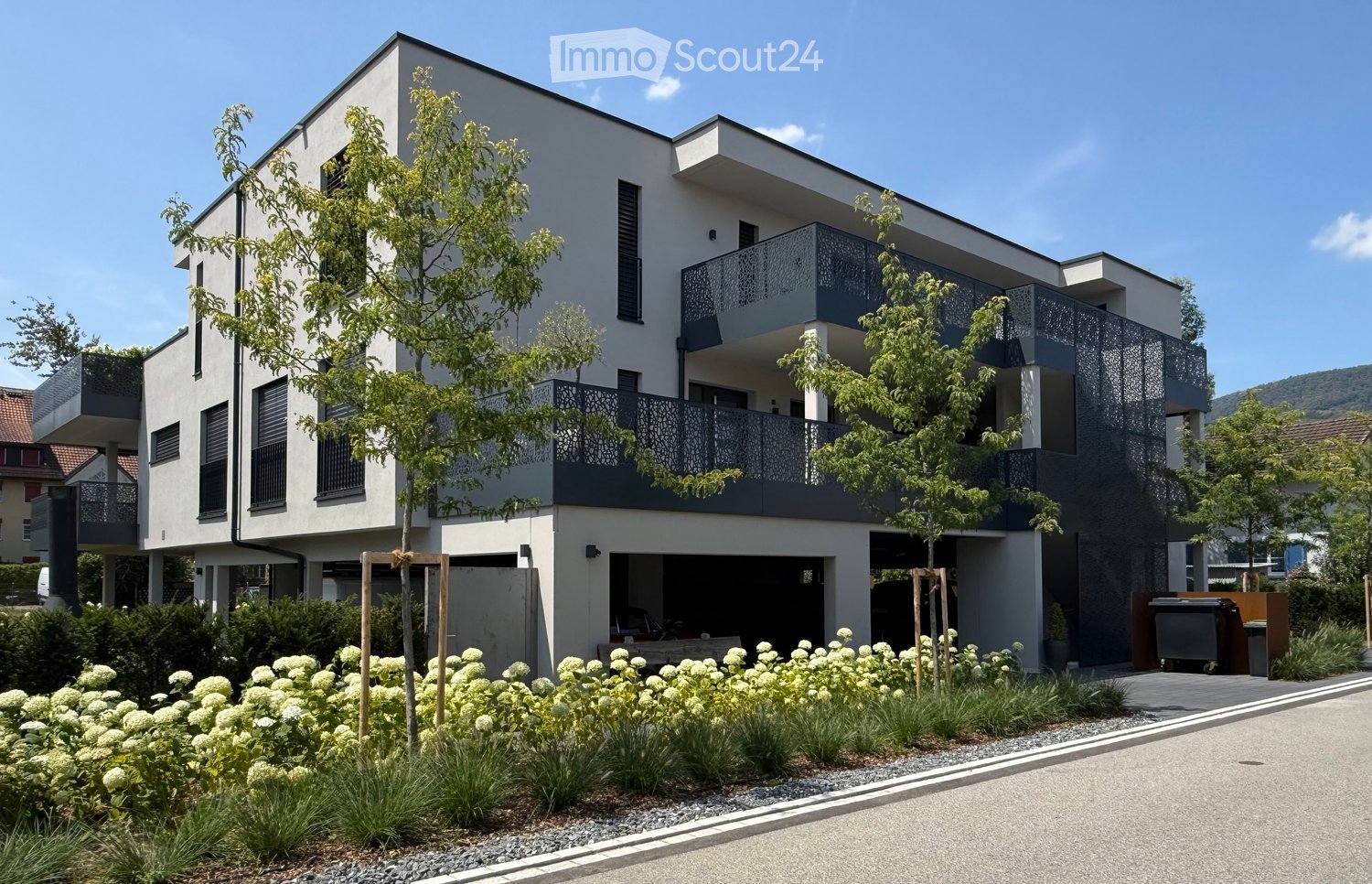 Modern two-story residential building with white and grey facade, balconies, and landscaped garden area with shrubs and trees.