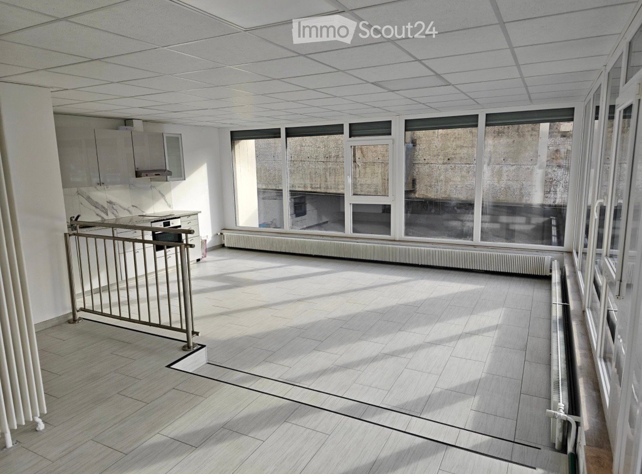 Empty room with tiled floor, white walls, large windows, and a metal railing. No furniture or equipment present.