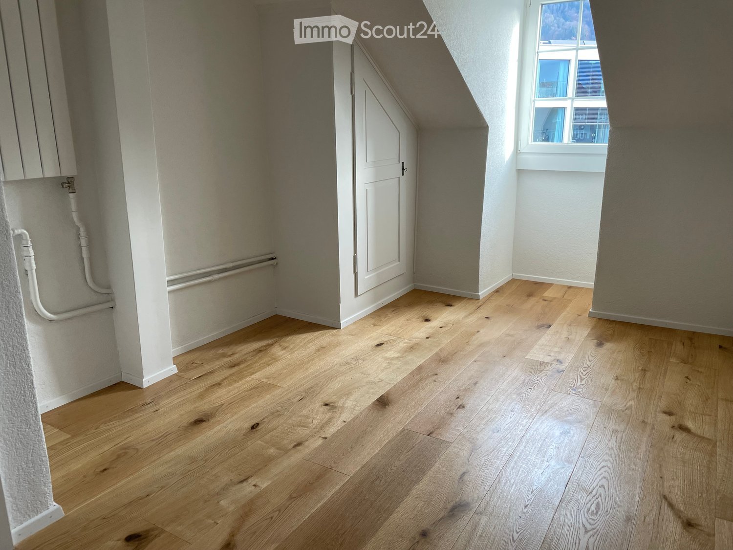 An empty attic room with wooden flooring, white walls, and a window letting in natural light. The room features a door and visible pipes along one wall.