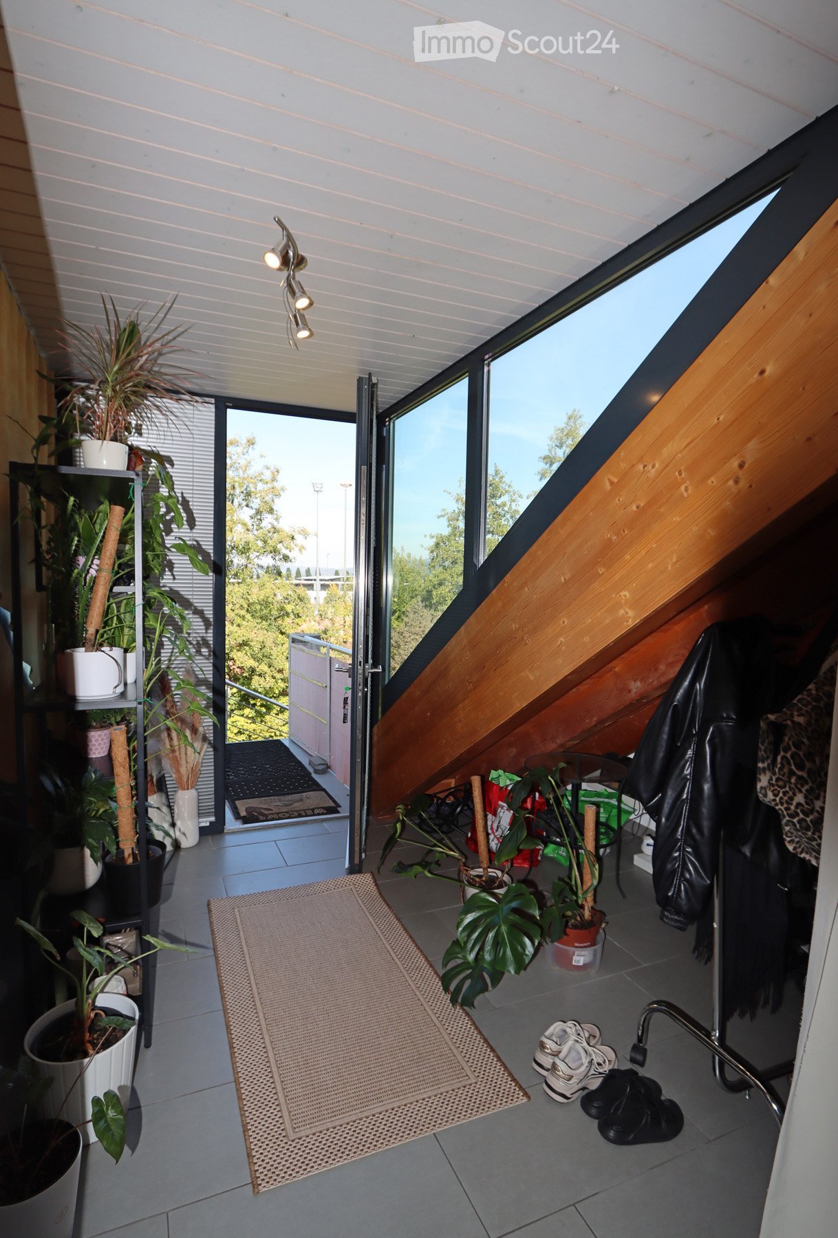 A modern, well-lit balcony with large windows and skylight, featuring potted plants, a woven rug, a small table, and a chair. Shoes and a coat are placed near the entrance.