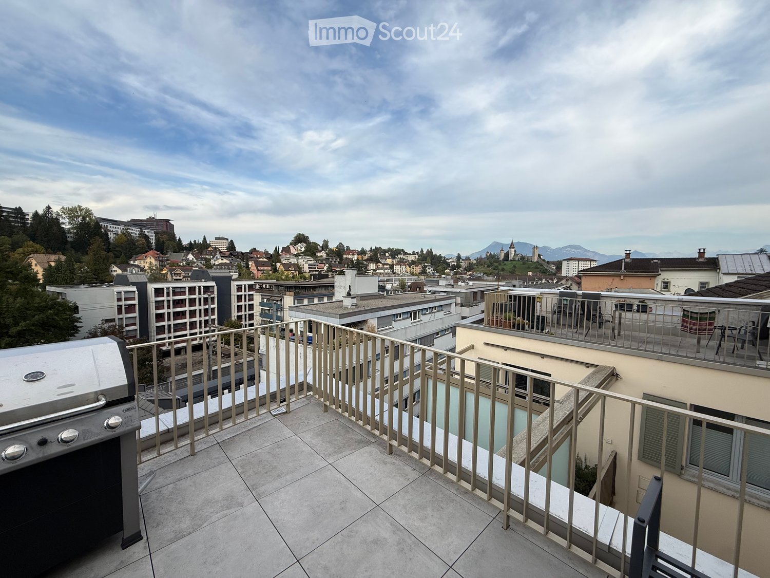 Balcony with a grill, overlooking a cityscape with buildings and mountains in the background.
