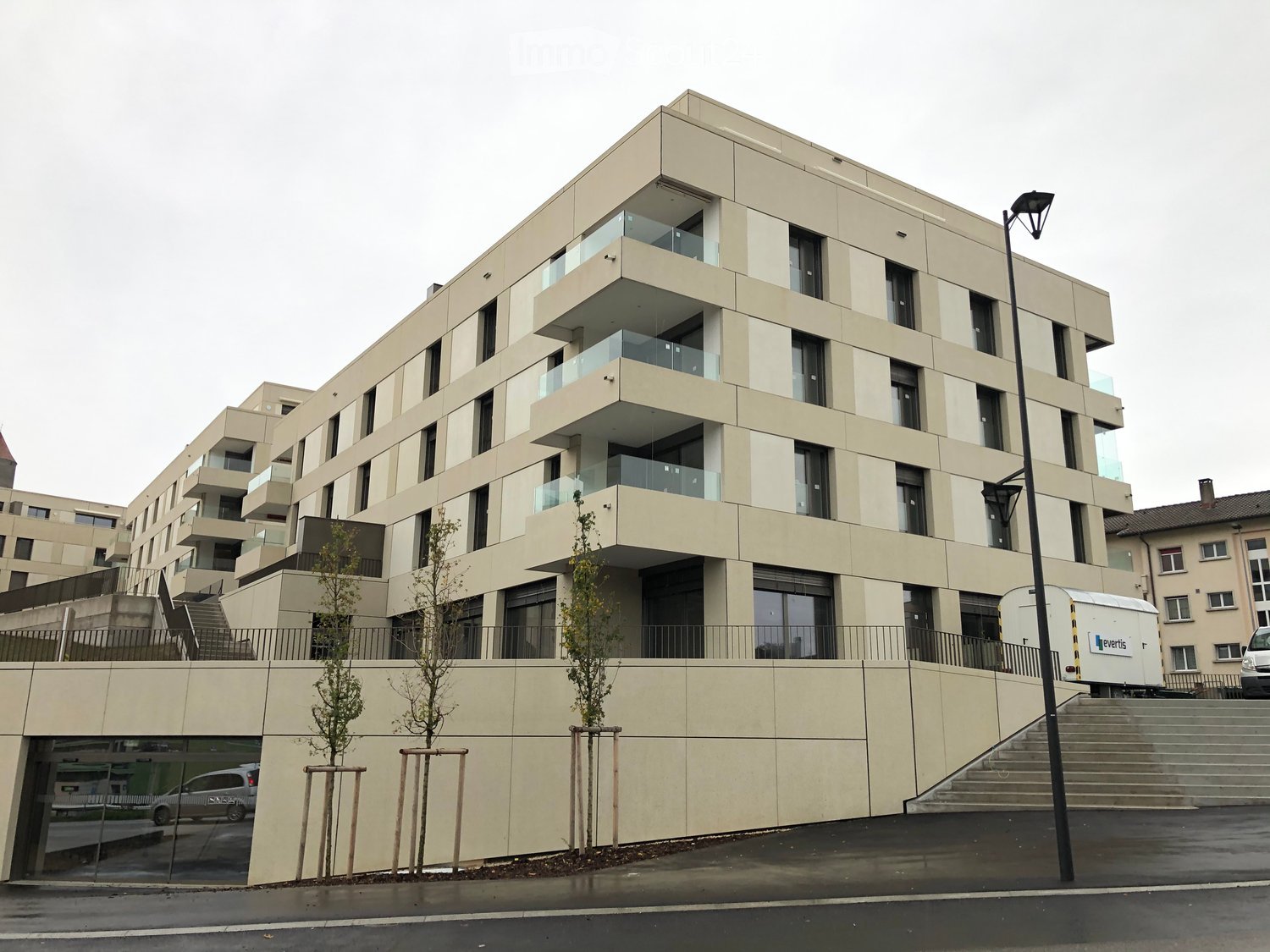 Modern multi-story residential building with beige facade, large windows, and balconies. Features a raised ground floor entrance and small landscaped area with young trees.