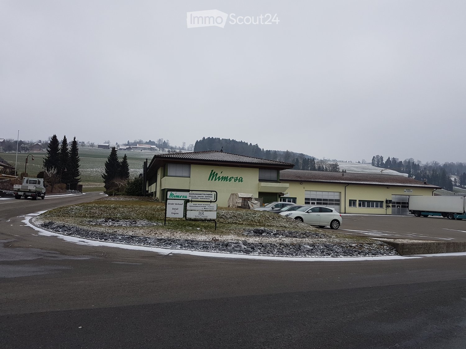 A two-story building with a yellow facade and a sign reading "Mimosa" in green letters. The building has multiple windows, a garage entrance, and a parking area with several cars and a truck. The surrounding area includes a gravel driveway, a grassy field