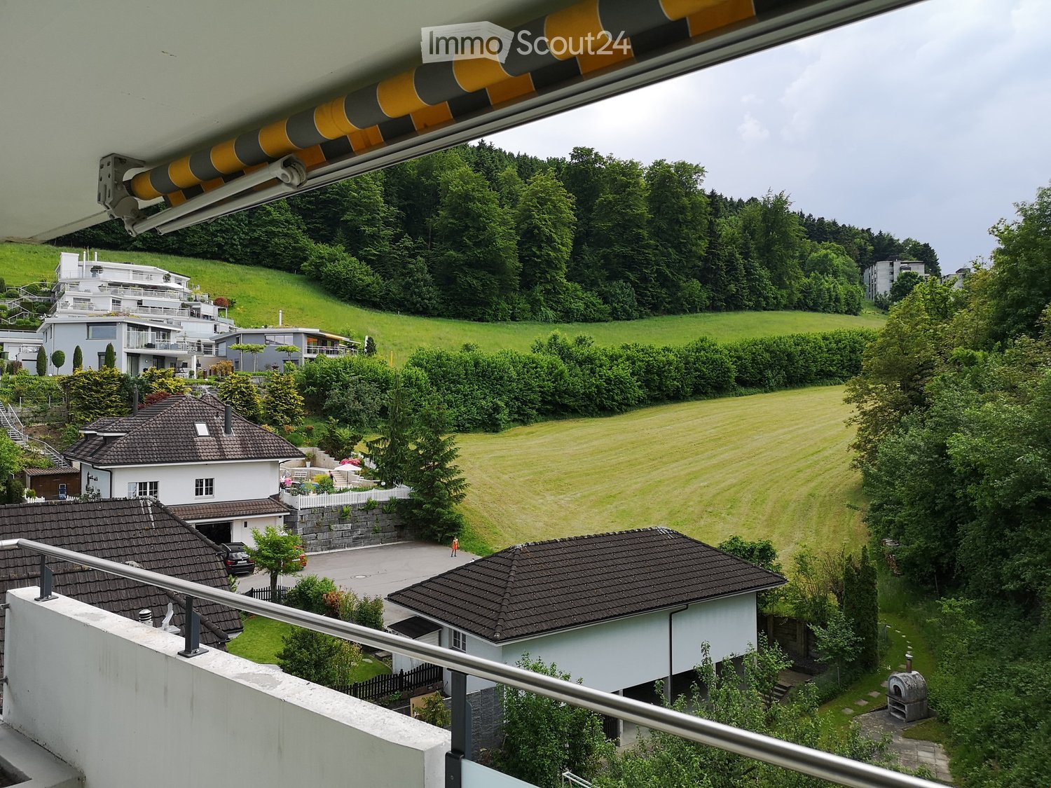 View from a balcony showing a green, hilly landscape with several houses, trees, and a well-maintained lawn.
