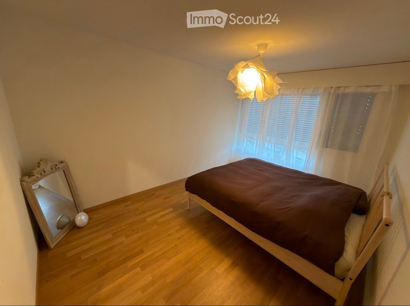 Cozy bedroom with wooden flooring, a single bed with dark bedding, a large mirror leaning against the wall, and a decorative hanging lamp.