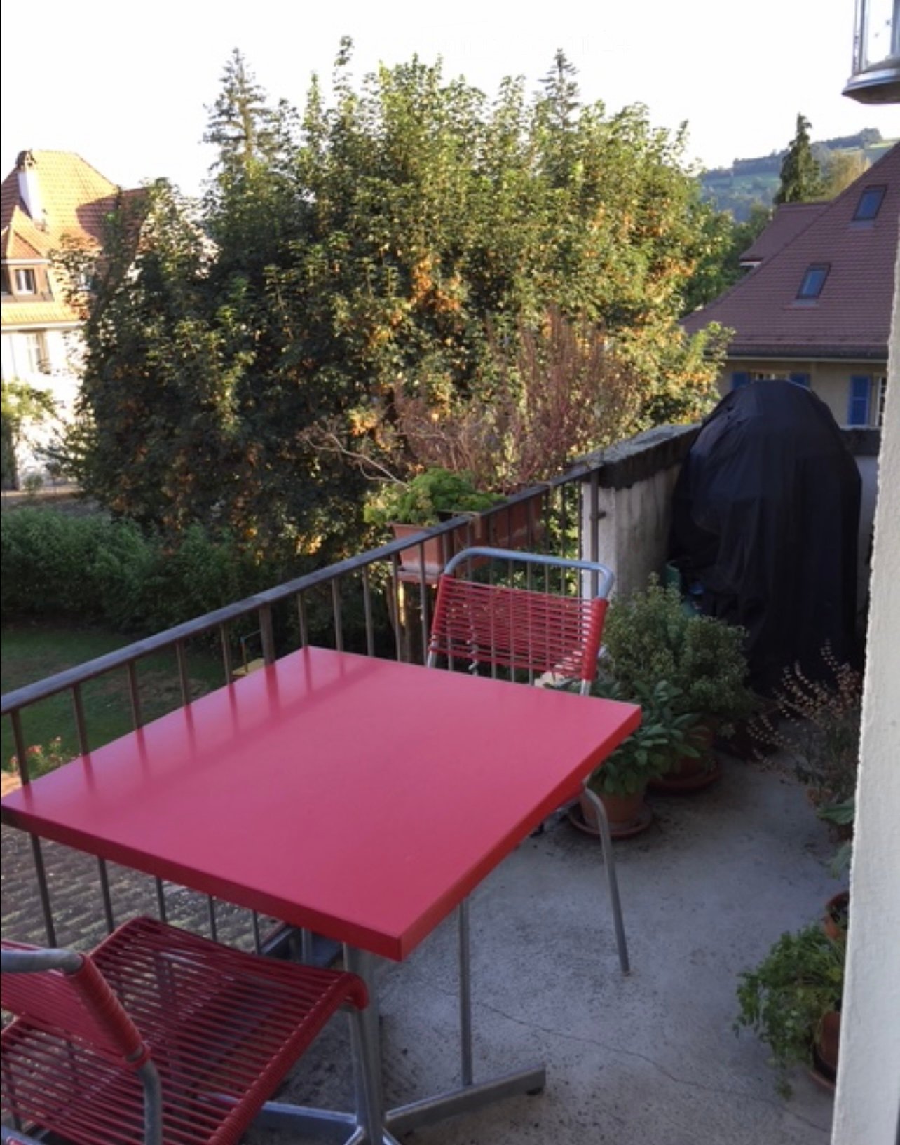 Balcony with a red table and matching red chairs, surrounded by potted plants and a covered grill, overlooking a green, tree-filled area.