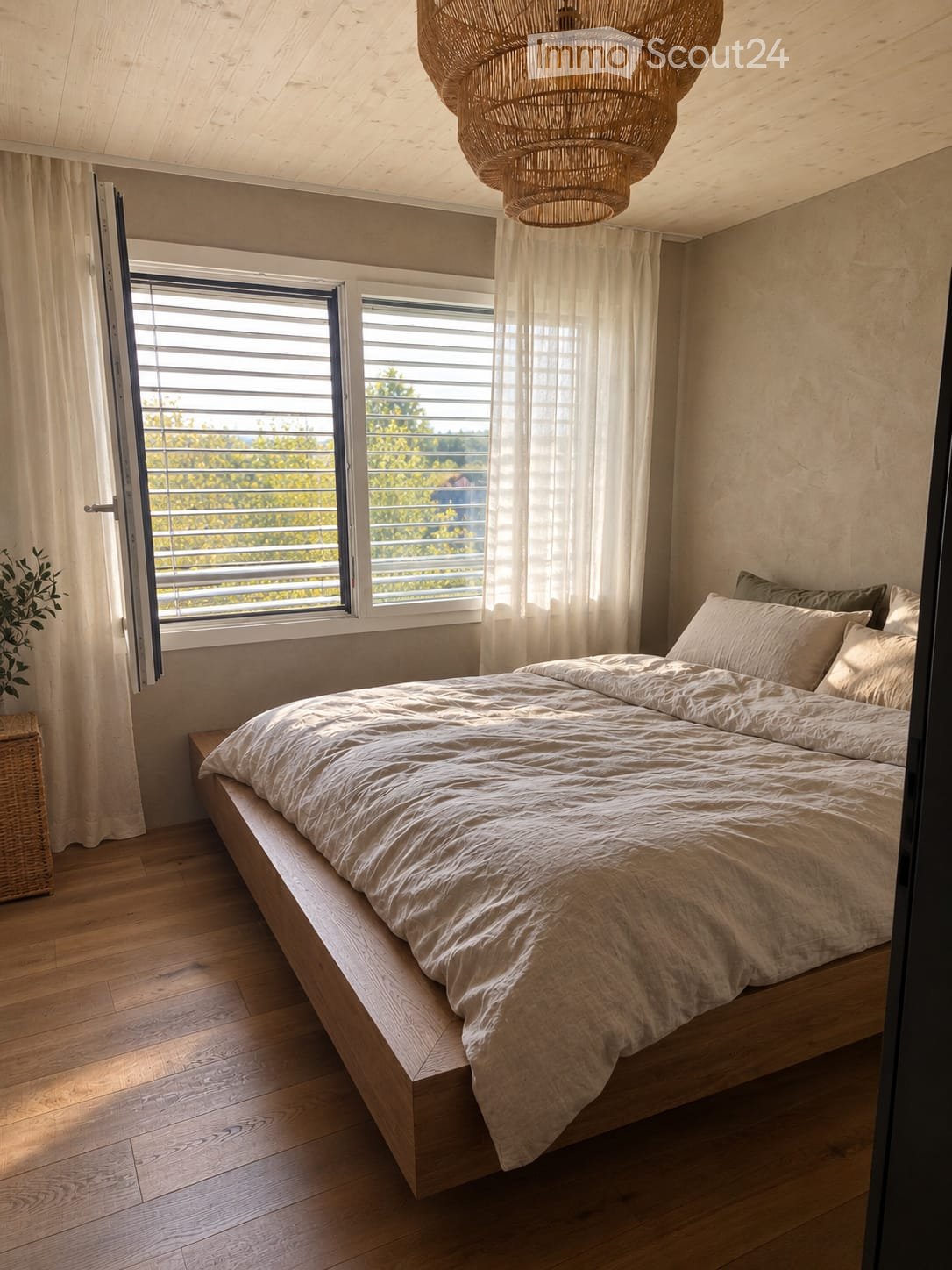 Minimalist bedroom with a wooden bed, neutral bedding, large windows with blinds, and a wicker chandelier.