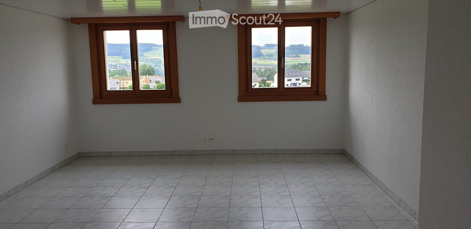 Empty room with light grey walls, tiled floor, and two large windows offering a view of the countryside.