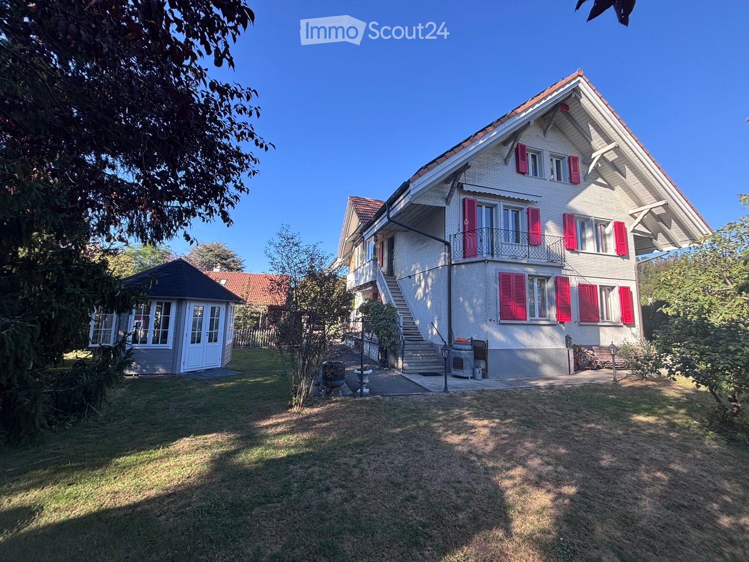 Two-story house with white exterior, red shutters, and a balcony. The house has a well-maintained lawn and a small shed in the background.