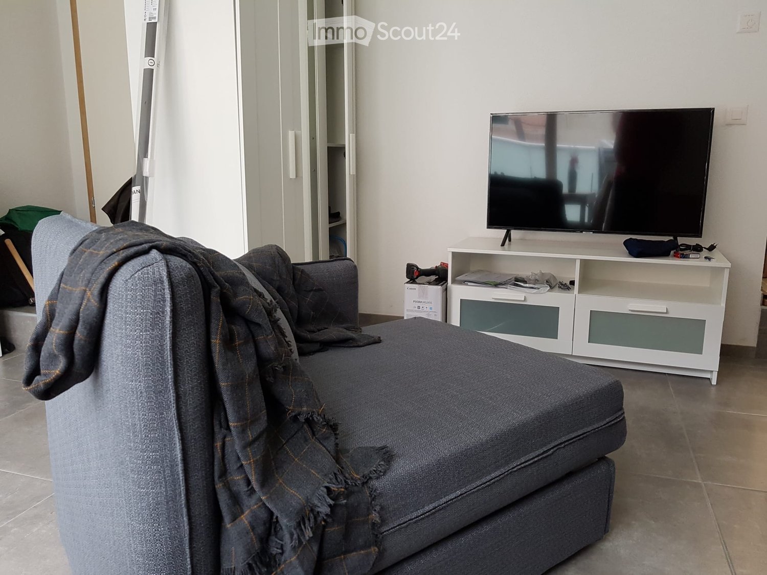 Living room with a grey sofa draped with a blanket, a flat-screen TV on a white cabinet, and a closet in the background.