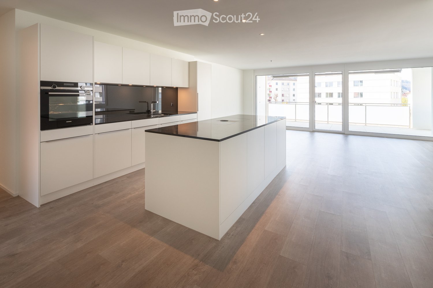 Modern kitchen with white cabinets, a central island, and a built-in oven. The space features wooden flooring and large windows offering a view of the city.