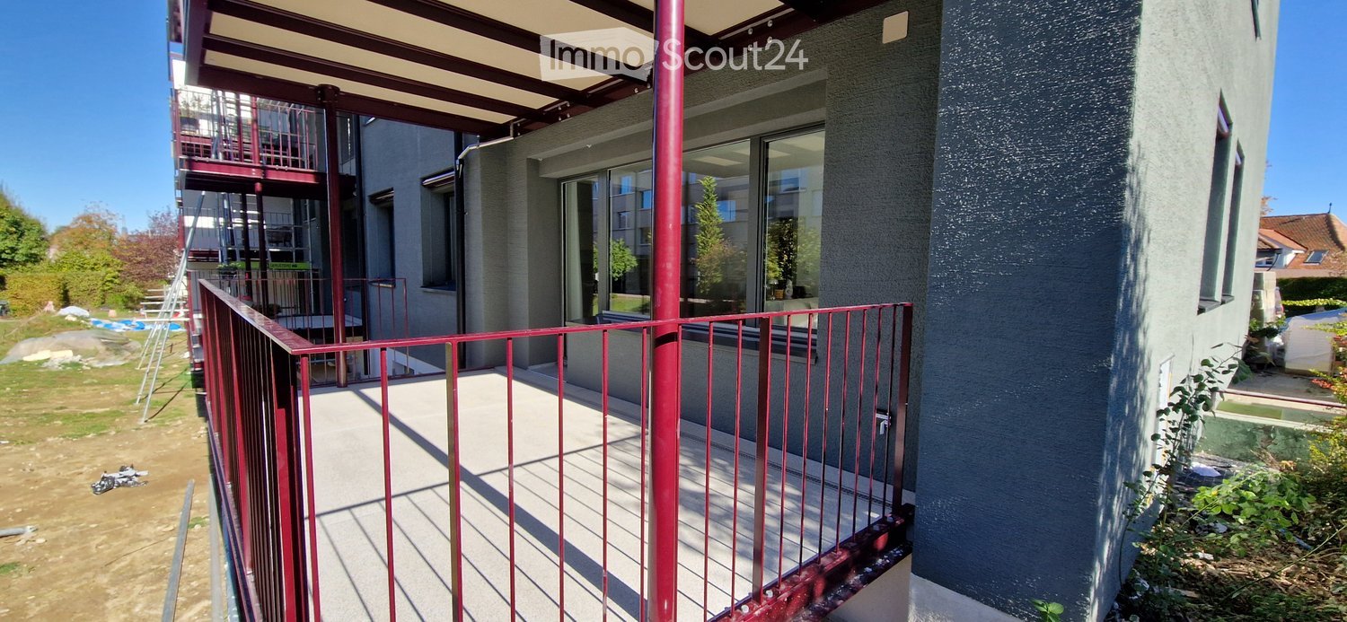 Balcony with red metal railing, concrete floor, and large windows overlooking the building.