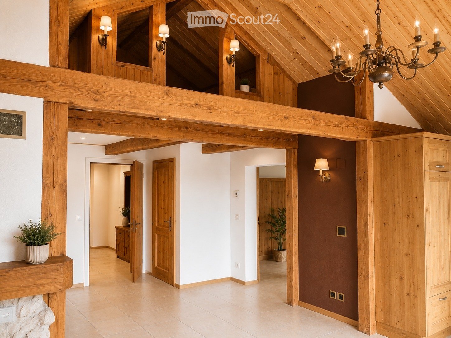 Spacious hallway with exposed wooden beams, warm lighting, and multiple doors leading to different rooms. Features a chandelier, potted plants, and a stone fireplace.