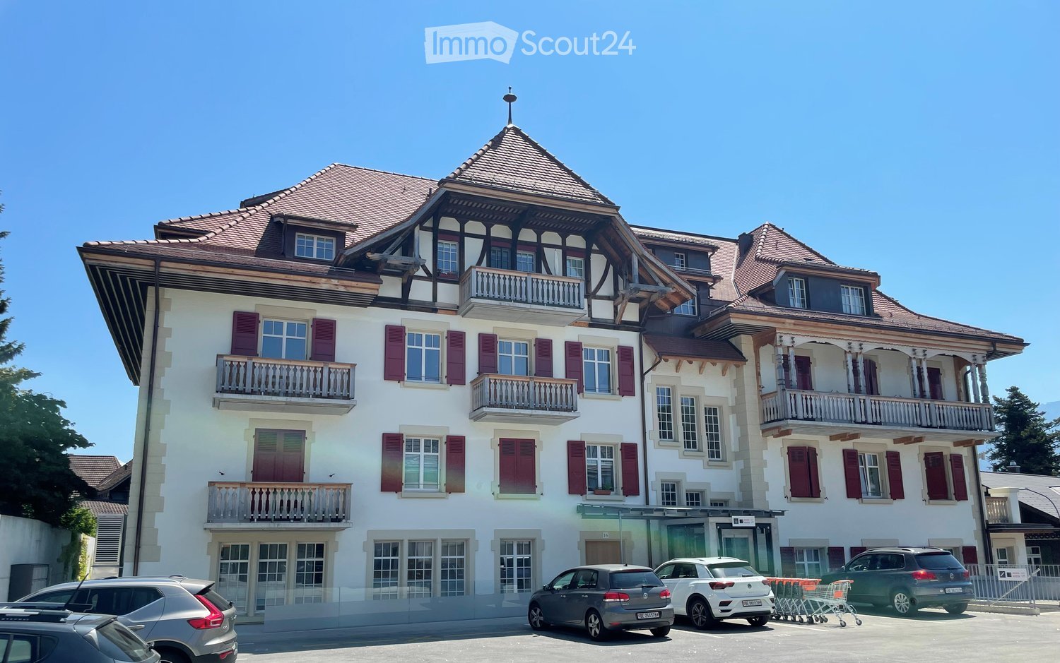 A large, multi-story building with white walls, red shutters, and multiple balconies. The roof is tiled with a distinctive wooden gable. Several cars are parked in front of the building.