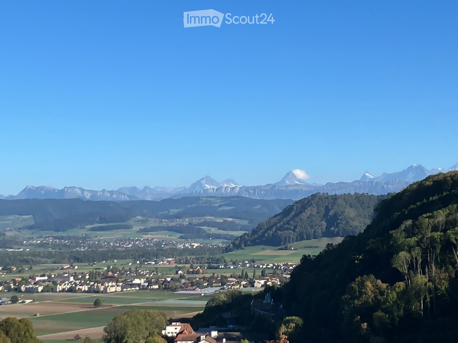 Panoramic view of a town nestled in a valley with surrounding mountains, green fields, and scattered houses.