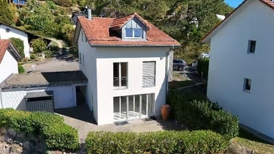 A modern two-story house with a red-tiled roof, white exterior, and large windows. The house features a small balcony and is surrounded by neatly trimmed hedges and a small garden area.