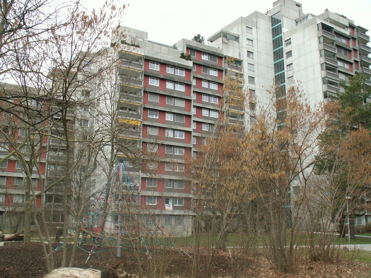 Multi-story residential building with red and grey facade, multiple windows, balconies, and a playground area in front.