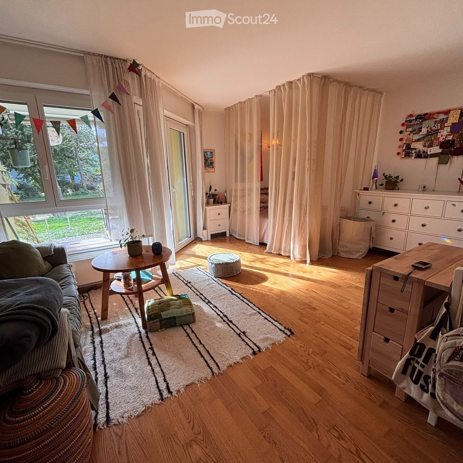 Bright living room with wooden flooring, large windows, and a sliding glass door leading to a garden. Features a grey sofa, small round table, colorful rug, and a wooden desk with a laptop.