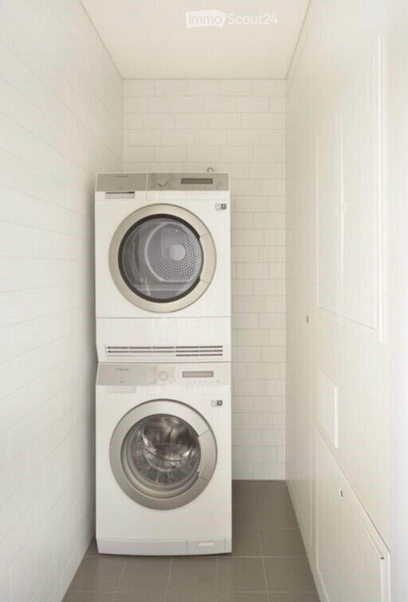 A small, modern laundry room with white tiled walls and a tiled floor, featuring a stacked washer and dryer unit.