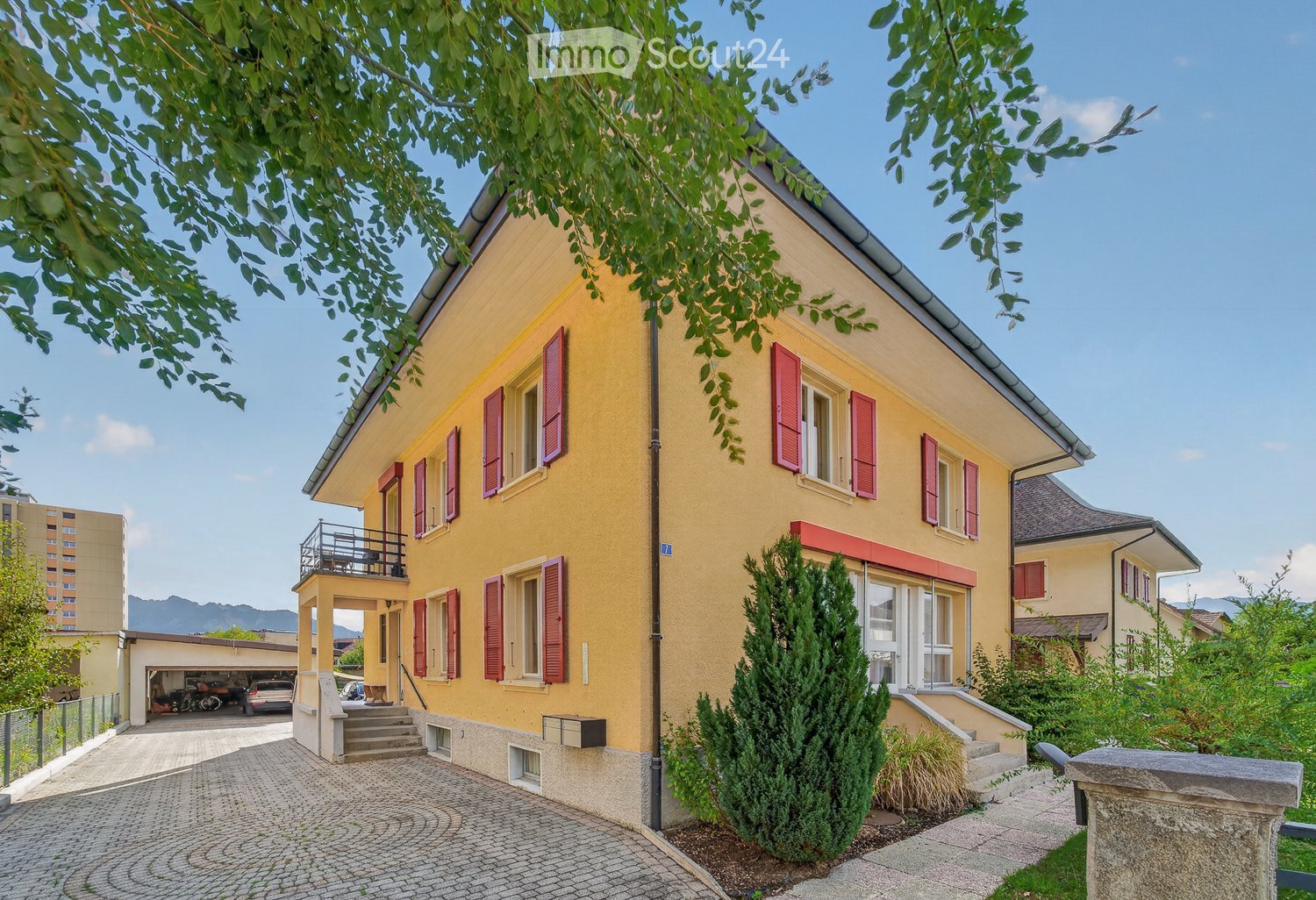 Two-story yellow house with red shutters, a balcony, and a garage. The house is surrounded by greenery and has a cobblestone driveway.
