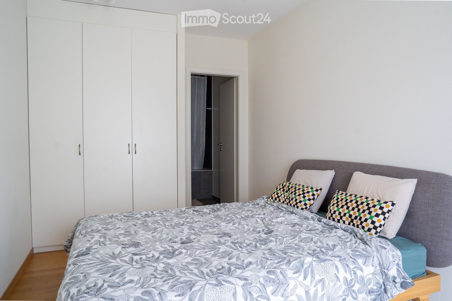 A modern bedroom with a grey upholstered bed, patterned pillows, a matching duvet, and a large white wardrobe. The room features wooden flooring and a visible doorway leading to another space.