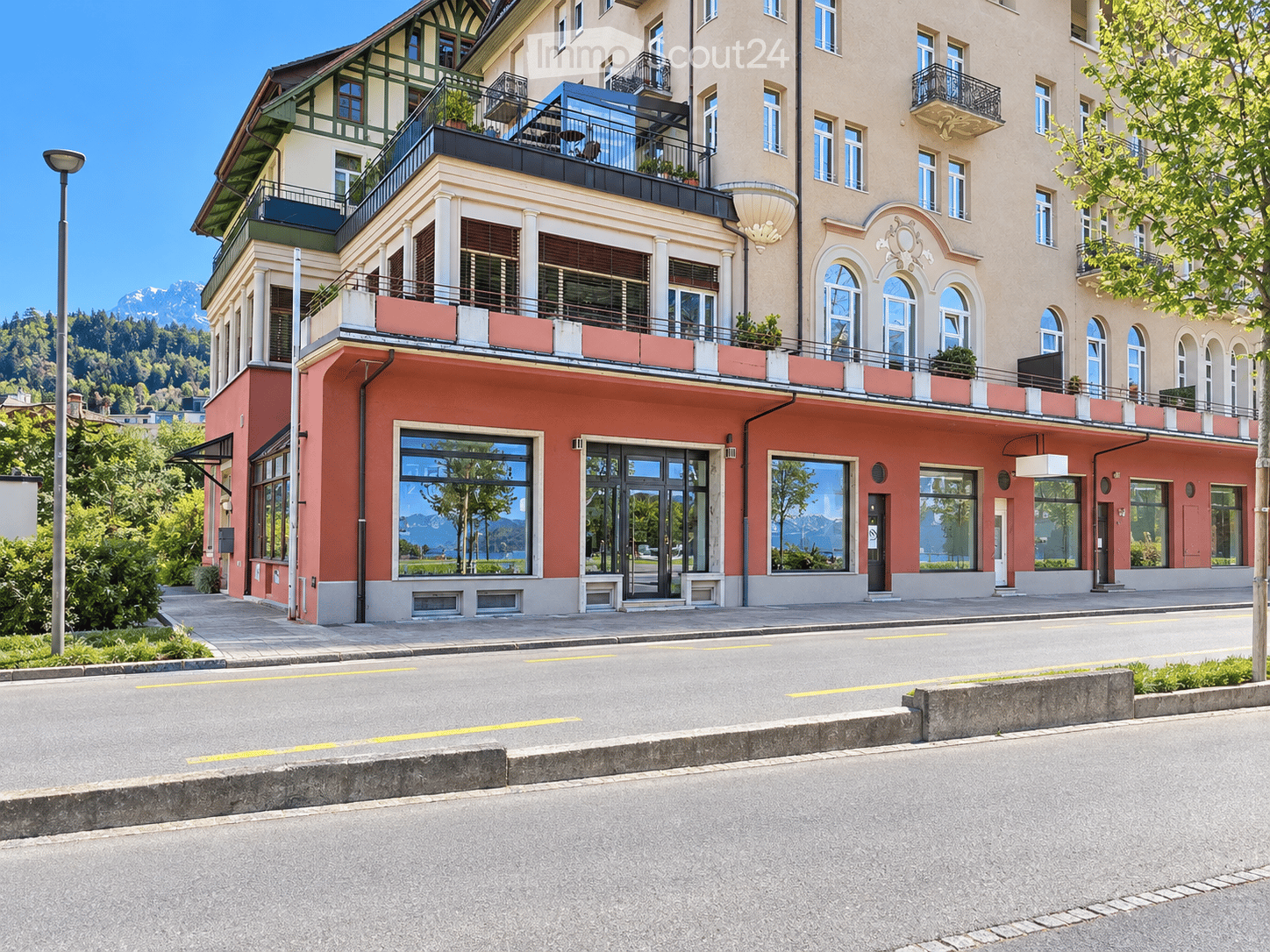 A multi-story building with a mix of modern and traditional architectural elements, featuring balconies, large windows, and a view of mountains in the background.