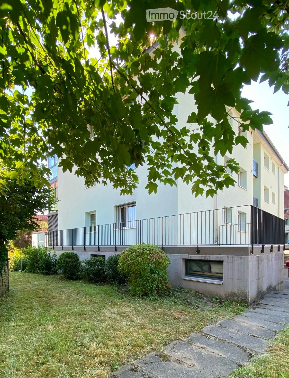 A well-maintained garden area with a modern two-story house in the background, featuring a balcony with metal railings, neatly trimmed bushes, and a stone pathway leading to the entrance.