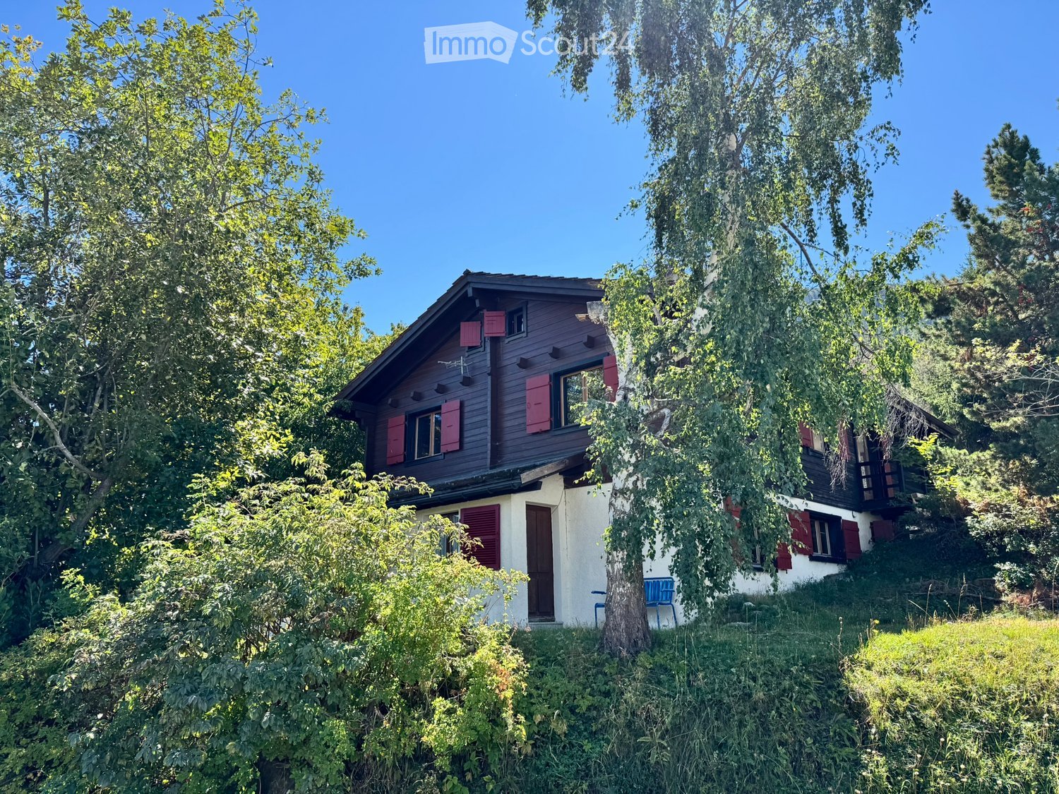 A two-story wooden house with red shutters and a sloped roof, surrounded by lush greenery and trees.