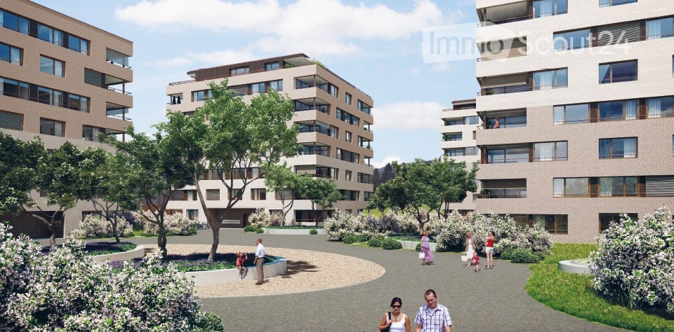 A modern residential complex with multiple apartment buildings, surrounded by landscaped gardens and pathways. The buildings feature contemporary architecture with large windows and balconies. The area includes trees, shrubs, and a central paved walkway w