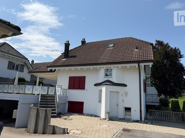 Two-story house with a brown tiled roof, white exterior, red shutters, and a small balcony on the left side. A garage is visible on the left, and the house is surrounded by a paved driveway and greenery.