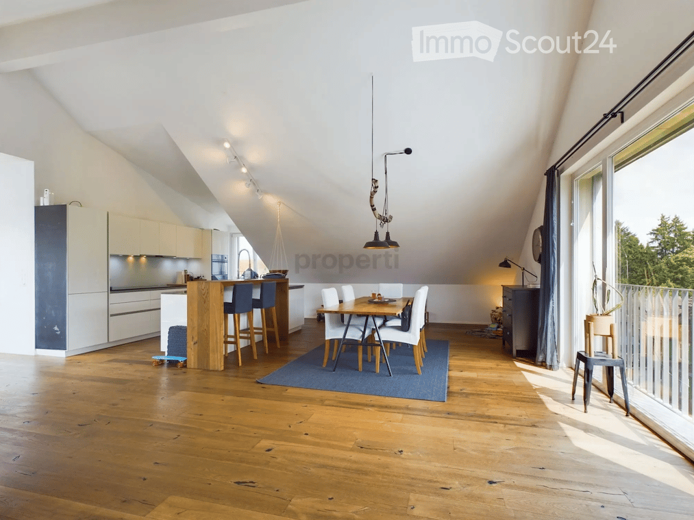 Open-plan kitchen and dining area with modern white cabinets, a central island, bar stools, a dining table with chairs, and a hanging pendant light. The space features wooden flooring and large windows letting in natural light.