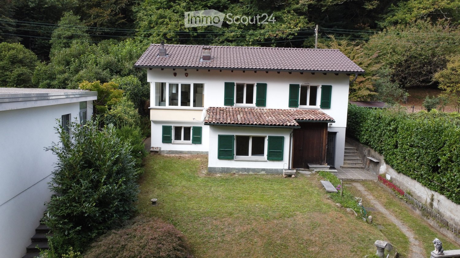 Two-story house with white walls, green shutters, and a tiled roof, surrounded by greenery and a small garden area.