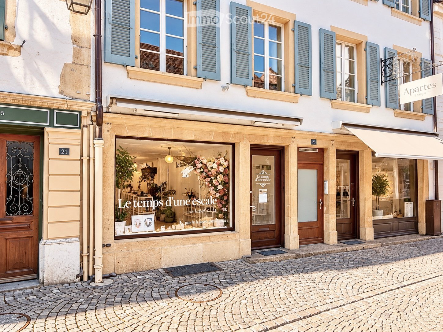 A charming multi-story building with classic European architecture, featuring light-colored stone facades, blue shutters, and multiple storefronts including a flower shop with visible floral arrangements and decorative items inside.