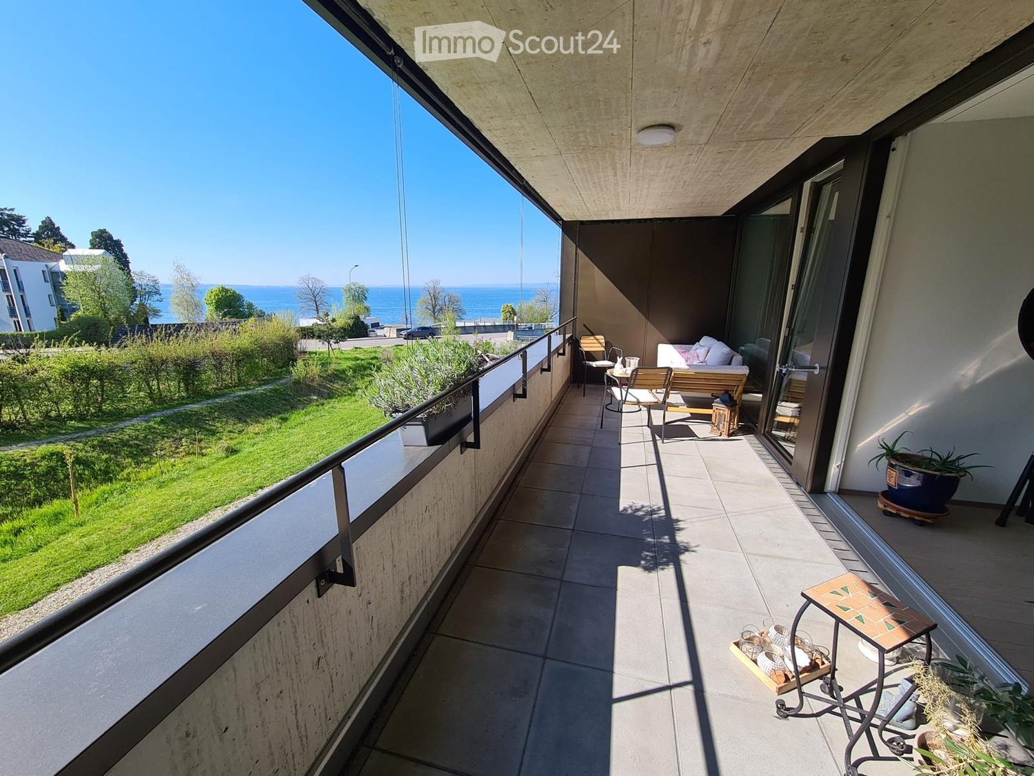 Balcony with tiled floor, modern railing, sofa, chairs, small table, and potted plants, overlooking a green lawn and sea view.