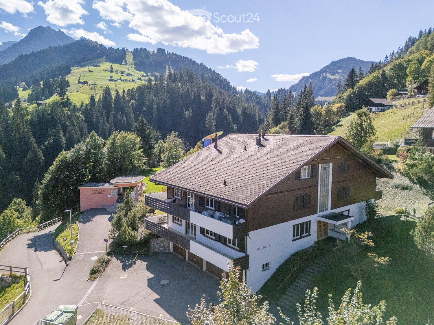 A two-story house with a brown roof and white walls, situated in a scenic mountainous area with a driveway and surrounding greenery.