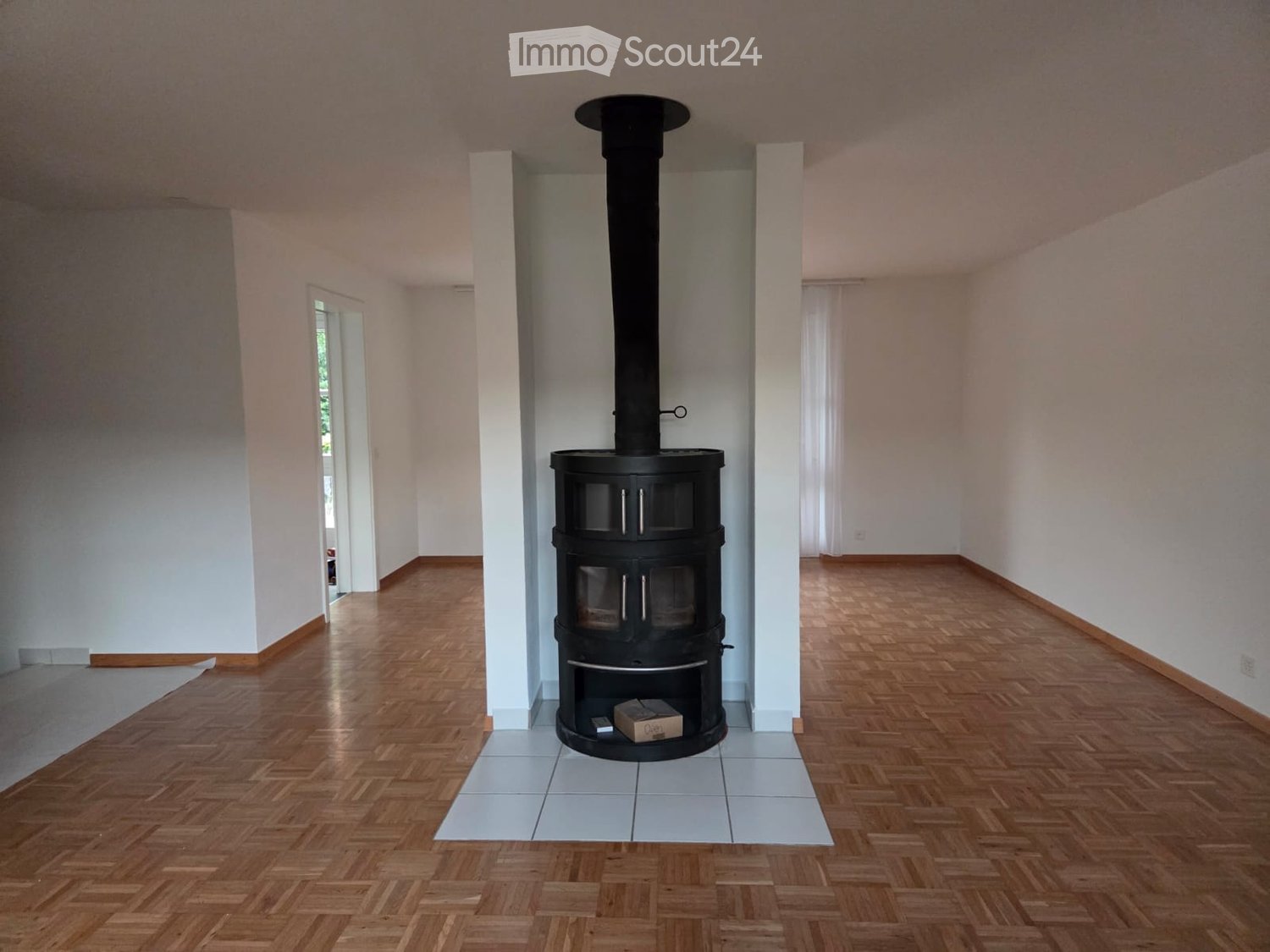 Empty room with wooden flooring, white walls, and a central black wood-burning stove with a chimney. The room has a tiled area around the stove and a doorway leading to another space.