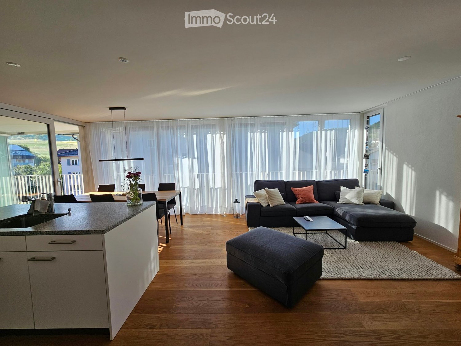 Open-plan living and dining area with a grey sectional sofa, coffee table, ottoman, dining table with chairs, and a kitchen island with a sink.