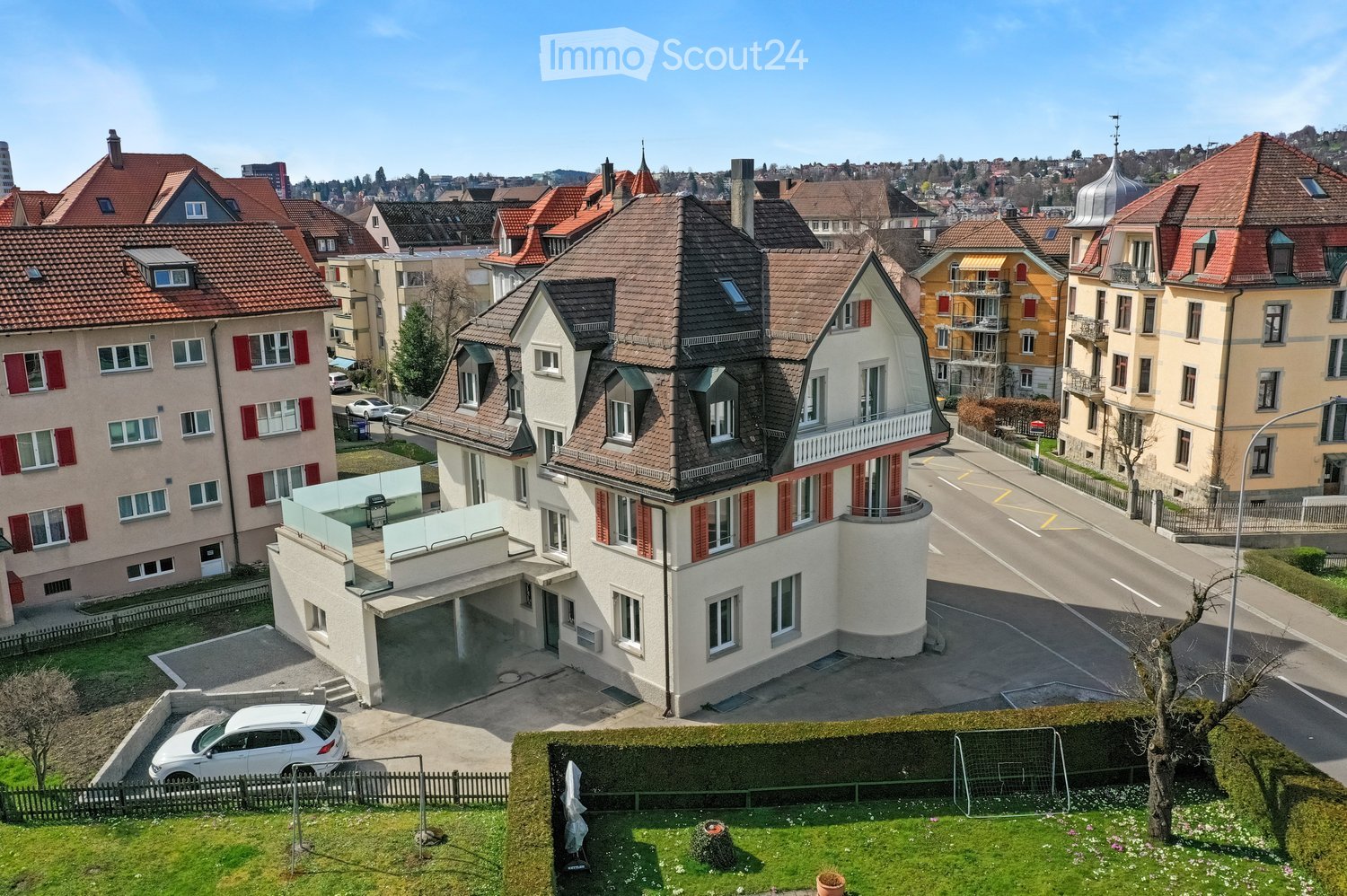 A multi-story house with a red-tiled roof, white walls, and red shutters. The house features a modern garage and a balcony on the upper floor. The surrounding area includes other residential buildings, a driveway, and a small garden with a tree and a socc