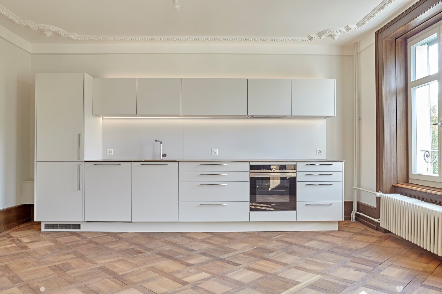 Modern kitchen with white cabinets, stainless steel appliances, and wooden herringbone flooring.