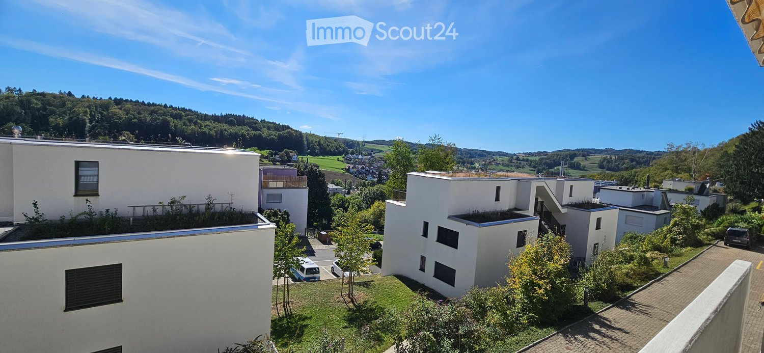 Modern residential complex with white buildings, green roofs, and surrounding greenery. Several parked cars and a scenic view of hills in the background.