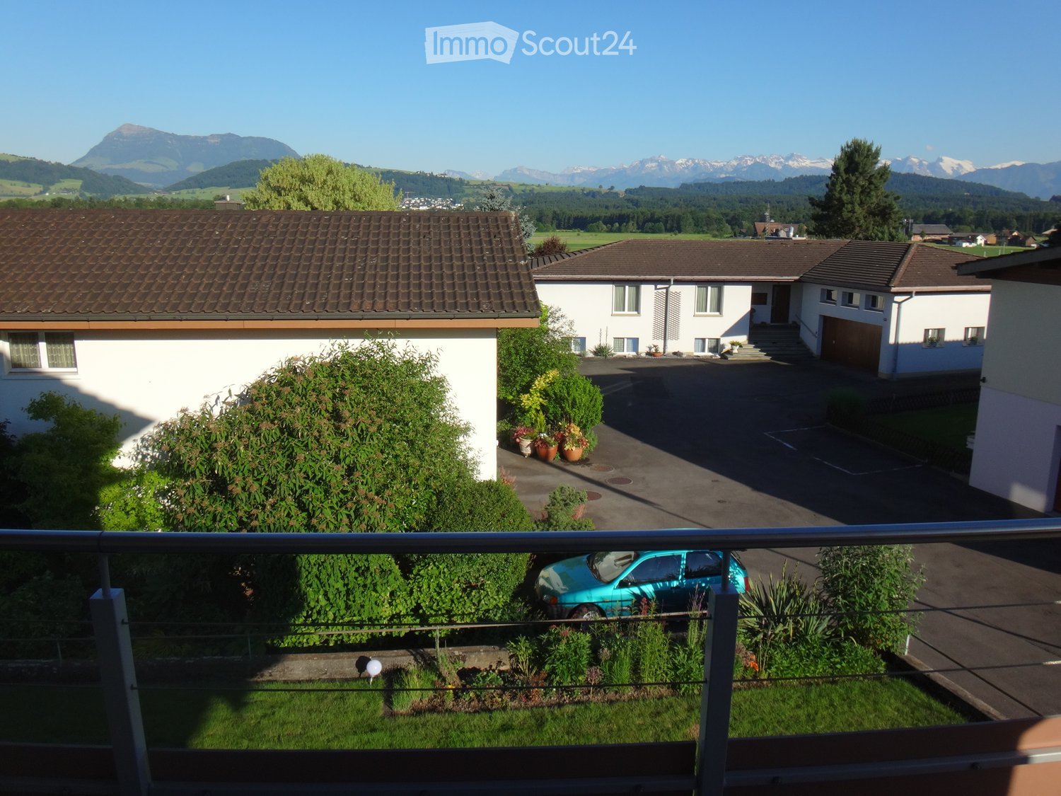Three houses with brown roofs, white walls, and a parking lot in front. A blue car is parked in the lot. The houses are surrounded by plants and trees. In the distance, there are mountains and a clear blue sky.