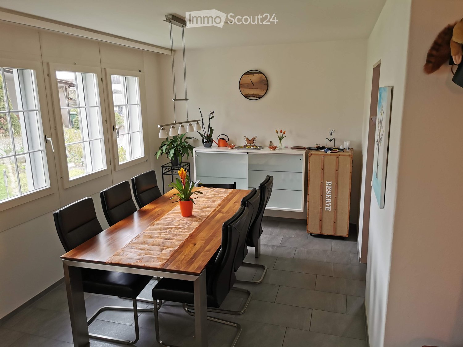 Bright kitchen with a wooden dining table surrounded by black chairs, a potted plant centerpiece, and a modern cabinet with glass doors. Large windows provide natural light.