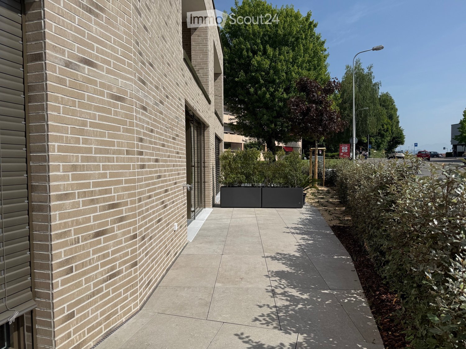 Modern building exterior with beige brick facade, large glass windows, and a paved walkway lined with greenery and shrubs.